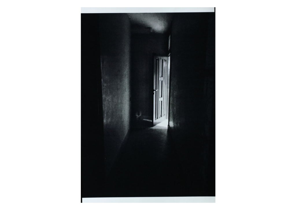 A black and white photo shows an open door at the end of a dark corridor. There is light coming in through the opening.
